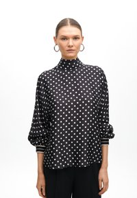 Schwarze Bluse mit Punkten, hohem Kragen, Ballonärmeln und weißen Akzenten an den Manschetten. Glatter Stoff mit leichtem Glanz.