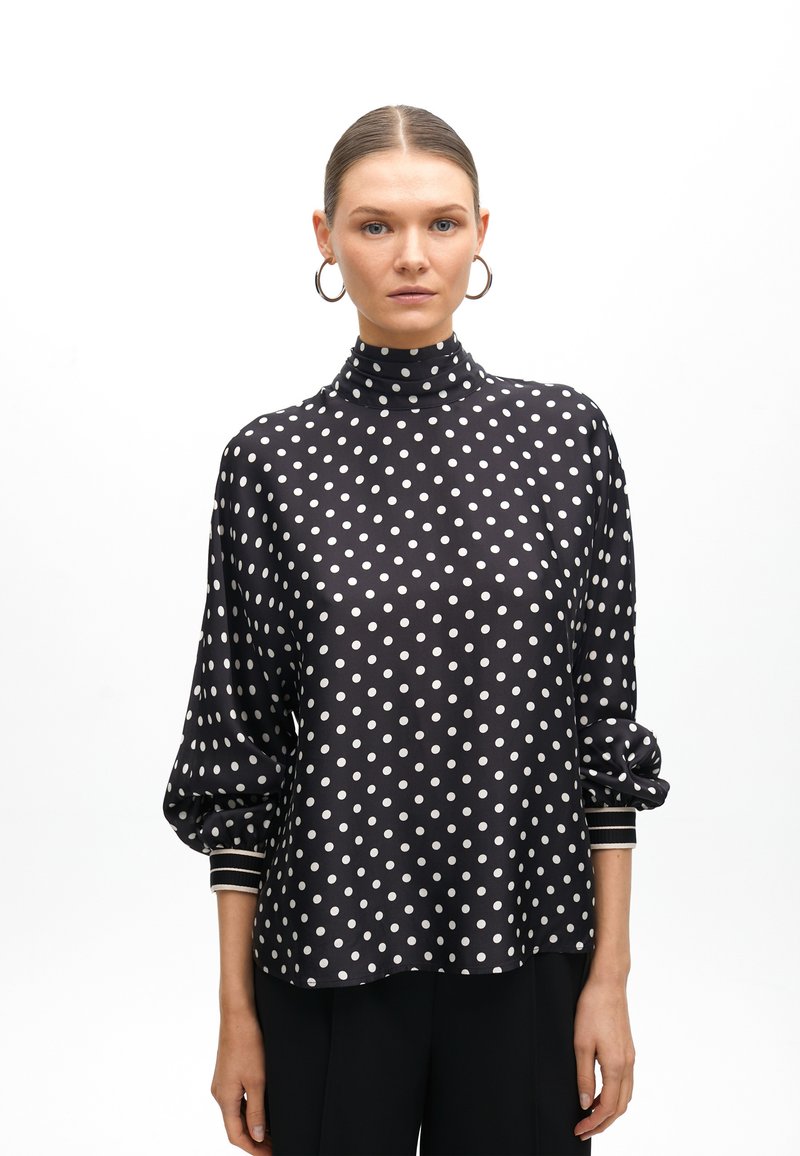 Schwarze Bluse mit Punkten, hohem Kragen, Ballonärmeln und weißen Akzenten an den Manschetten. Glatter Stoff mit leichtem Glanz.