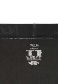 Zwarte stof met een gladde textuur, bevat het verzorgingslabel en maatdetails. Versterkte stiksels langs de bovenrand, mernaam gedrukt in donkergrijs.