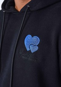Sudadera negra con dos corazones azules bordados, uno más grande que el otro, encima del texto "Project & Paris HEART STUDIO."