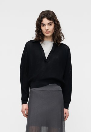 Jonge vrouw met krullend haar, gekleed in een zwarte V-hals trui over een licht topje en een doorschijnende grijze rok, staand tegen een effen achtergrond.