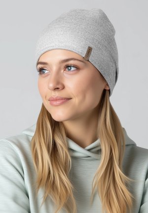 Grijze gebreide muts met een zachte textuur, een afgeronde vorm en een klein bruin stoffen label aan de zijkant; gedragen met een lichtgroene hoodie.