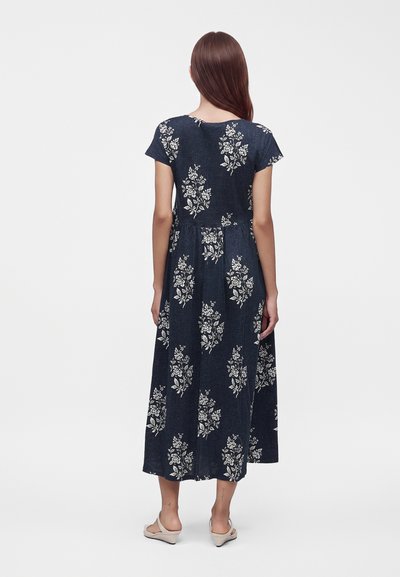 Femme aux longs cheveux bruns portant une robe midi bleu marine à manches courtes imprimée de fleurs et des chaussures plates beiges, debout de dos.
