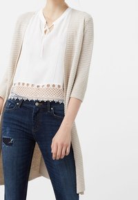 Beige pletený cardigan, bílý top s vertikálními sklady a krajkovým lemem, tmavě modré džíny se záplatu, ruka modelky opřená o bok.