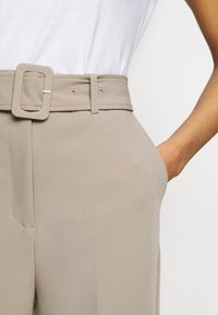 Pantalons taille haute beige clair avec une large ceinture à boucle carrée, poches latérales et tissu lisse et texturé.