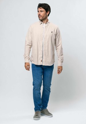 Mann trägt beige Hemd mit Knöpfen, weißes Unterhemd, blaue Jeans und graue Turnschuhe, steht und schaut nach links vor einem schlichten Hintergrund.