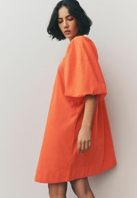 Pomarańczowa lniana sukienka o luźnym kroju, z krótkimi puffowymi rękawami i długości do kolan. Teksturowana tkanina ma miękki, lekki wygląd.