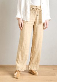 Beige linen wide-leg pants with a drawstring waist and small metallic accents, paired with tan shoes featuring a textured design.
