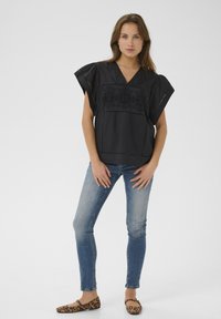 Fekete, rövid ujjas blúz hímzett részletekkel és V-kivágással, amelyet testhezálló kék farmernadrággal és leopárdmintás lapos cipővel viselnek.