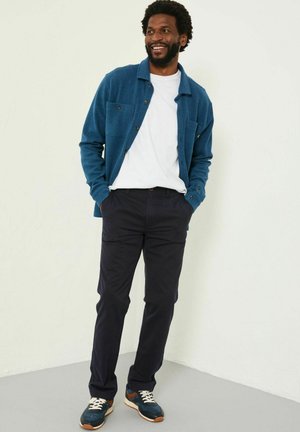 Blau gemustertes Button-Up-Hemd, weißes T-Shirt, marineblaue Hose und Sneakers mit blauen und braunen Akzenten bilden ein lässiges Outfit.