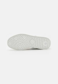 Semelle d'une basket blanche avec des motifs texturés et des adhérences circulaires sur un fond clair uni.