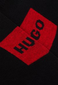 Tissu en tricot noir avec un carré rouge portant le mot "HUGO" en lettres noires en gras, soulignant un design moderne et une texture.