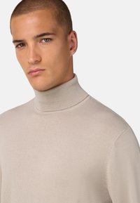 Bež pulover s turtleneck ovratnikom, izdelan iz rebraste pletenine, ki se ponaša z visokim ovratnikom in prilegajočo silhueto. Gladka tekstura brez vidnih vzorcev.