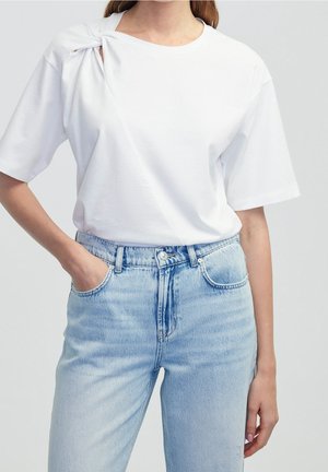 Femme portant une chemise blanche à manches courtes avec un nœud à l'épaule et un jean bleu clair taille haute, les mains dans les poches.