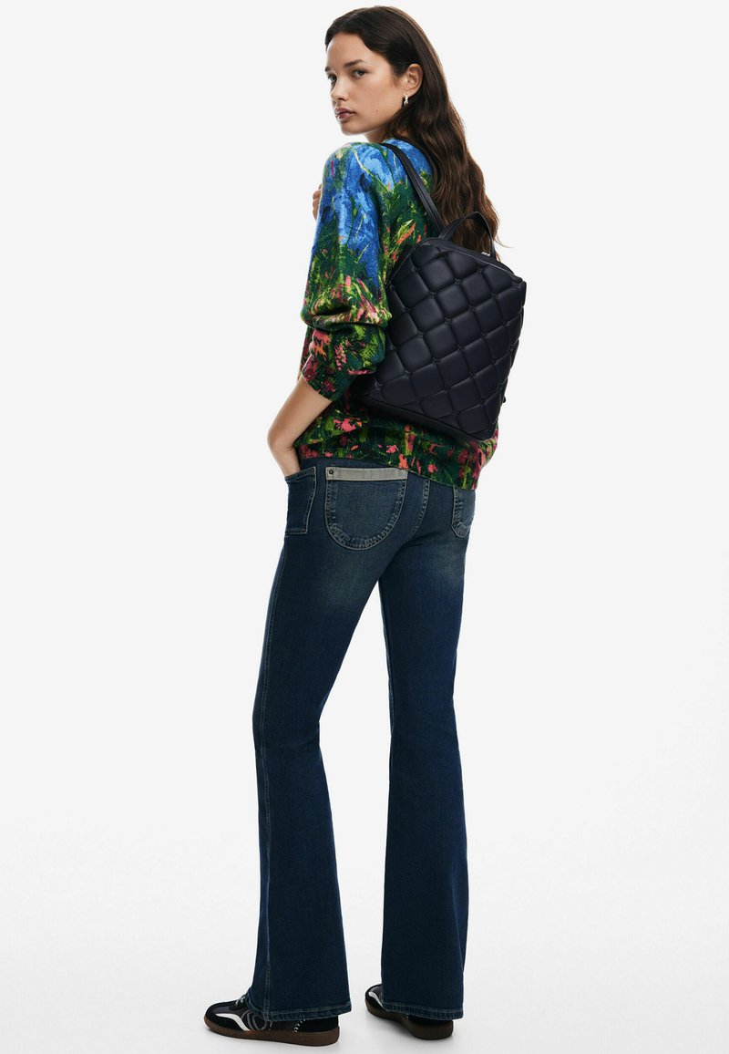 Czarna pikowana plecak z dwoma ramiączkami, noszona na kolorowy wzorzysty sweter i ciemne dzwony, ukazująca gładką fakturę materiału.