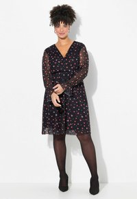 Schwarzes Kleid mit durchsichtigen Ärmeln, das ein Herzmuster in Rot und Rosa aufweist. V-Ausschnitt und elastischer Bund. Ergänzende schwarze Strumpfhosen und Schuhe.