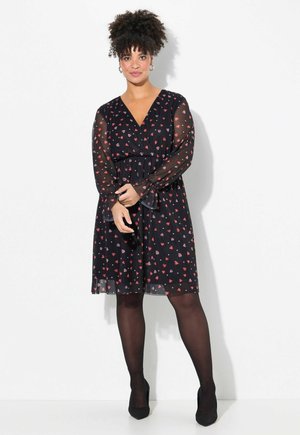 Zwarte jurk met doorzichtige mouwen, versierd met een hartpatroon in rood en roze. V-halslijn en elastische taille. Bijpassende zwarte panty en schoenen.