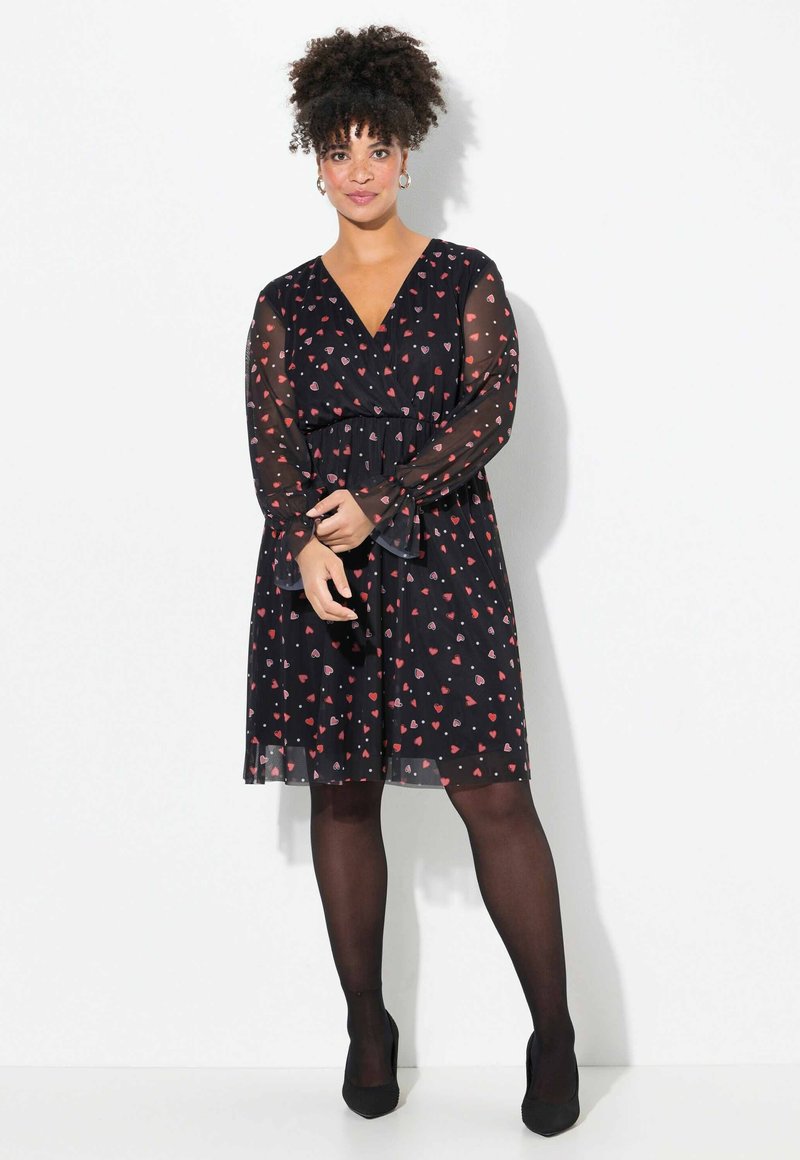Schwarzes Kleid mit durchsichtigen Ärmeln, das ein Herzmuster in Rot und Rosa aufweist. V-Ausschnitt und elastischer Bund. Ergänzende schwarze Strumpfhosen und Schuhe.