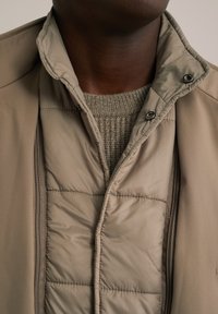 Chaqueta acolchada en beige claro con cierre de cremallera y botones a presión. Presenta un suéter suave y texturizado debajo en un tono neutro complementario.