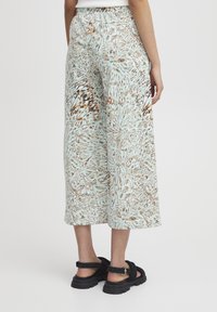 Pantalons large bleu clair avec un motif texturé marron et crème, dotés d'une taille haute et d'une coupe décontractée. Portés avec des sandales noires.