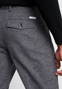 Grå rutiga byxor med en vävd struktur. Har en bakficka med knapp och en märkesetikett. Skräddarsydd passform med en klassisk design.