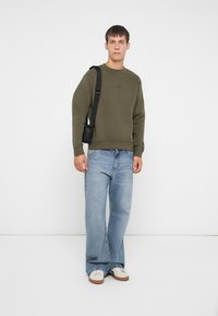 Oliwkowa bluza z okrągłym dekoltem i ściągaczami na rękawach, zestawiona z jasnoniebieskimi spodniami typu wide-leg i białymi sneakersami. Mała czarna torebka noszona na krzyż.