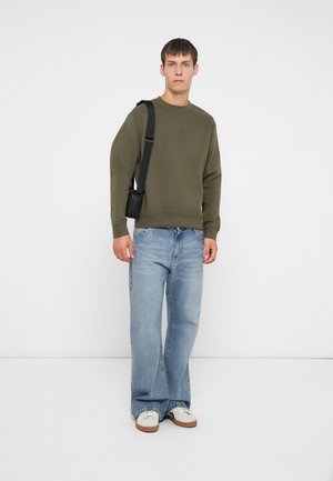 Sweat à col rond olive avec des poignets côtelés, associé à un jean ample bleu clair et des baskets blanches. Un petit sac noir est porté en bandoulière.