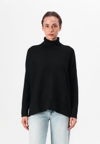 Pull à col montant noir avec une coupe ample, des épaules tombantes et une texture côtelée à l'ourlet et aux poignets ; associé à un jean bleu clair.