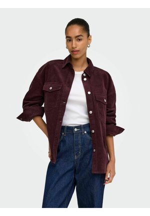 Junge Frau trägt eine bordeauxfarbene Cordjacke über einem weißen Hemd und blauen Jeans, steht mit einer Hand in der Tasche vor einem einfarbigen Hintergrund.