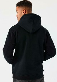 Hombre de perfil vistiendo un sudadera con capucha negra y con las manos en los bolsillos, contra un fondo claro y liso.