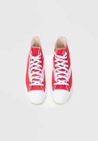 Raudonos aukštakulnės sportbačiai su baltais gumos pirštų dangteliais ir raišteliais, pagamintos iš drobės medžiagos su metalinėmis akutėmis, parodytos viršaus vaizde centre.