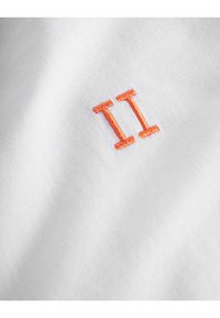 Hvit stoff med oransje broderte tall "II" sentrert på materialet, som viser en jevn tekstur og rent design.