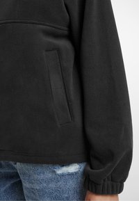 Svart fleece-pullover med sidelomme, ribbet mansjetter og myk tekstur, kombinert med slitte denimjeans.