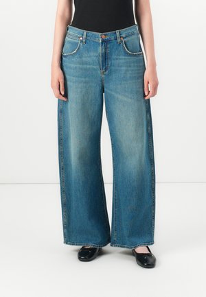 Persoon met loszittende blauwe denim jeans met zijstrepen, een zwart mouwloos topje en zwarte ballerina's, staand op een witte vloer.