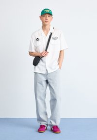 Camisa blanca de manga corta con parches, pantalones de mezclilla azul claro y zapatillas rosas. Una gorra verde completa el look, añadiendo color y estilo.