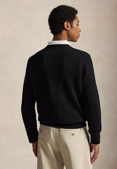 Hombre que lleva un jersey negro de punto sobre una camisa blanca con cuello y pantalones beige, de pie de espaldas con la cabeza girada hacia un lado.