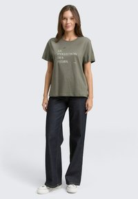 Olivgrünes Baumwoll-T-Shirt mit kurzen Ärmeln und weißem Schriftzug, kombiniert mit dunkelblauen Jeans mit weitem Bein und weißen Turnschuhen.