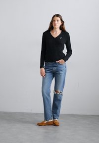 Black cable-knit sweater, v-neck, paired with light blue distressed jeans featuring frayed hem, and brown loafers. Plain gray background.
