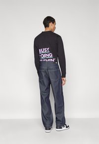 Camisa negra de manga larga con gran texto morado en la parte posterior, combinada con jeans de mezclilla oscuros y holgados, y zapatillas negras.