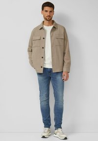 Beige blouse met knopen en twee voorzakken, gedragen over een lichtgrijs shirt, gecombineerd met een blauwe spijkerbroek en beige sneakers met patroon.