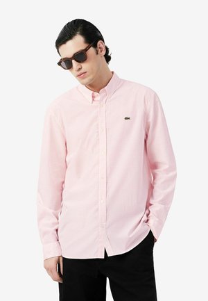 Hombre con gafas de sol marrones, camisa rosa claro con botones y pequeño logo, y pantalones negros, de pie con una mano en el bolsillo.