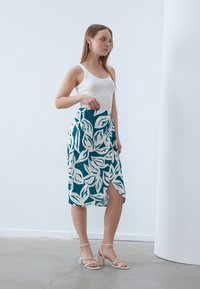 Teal skjørt med hvite bladmønstre, med en slitt på den ene siden. Glatt stoff, kne-lengde design, kombinert med lyse blokkhelte sandaler.