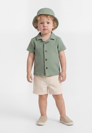 Kleinkind mit lockigem Haar, trägt ein grünes Hemd mit Knöpfen, passenden Hut, beige Shorts und beige Slipper, steht vor einem weißen Hintergrund.