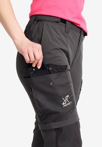 Person som bär mörkgrå byxor med sidoficka märkt RVRC, stoppar en svart smartphone i fickan, bär en rosa skjorta.