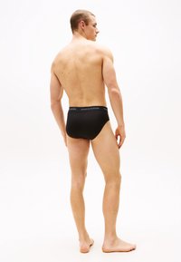 Schwarze enge Unterwäsche mit einem kontrastierenden, gebrandeten Bund. Das Modell steht barfuß, zeigt glatte Haut und einen neutralen Hintergrund.