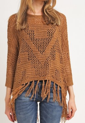 Žena nosící volný hnědý pletený svetr s třásněmi na lemu a modré džíny, stojící před jednobarevným pozadím.