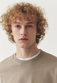 Felpa beige con scollatura a girocollo a coste, indossata sopra una t-shirt bianca. Il soggetto ha capelli ricci di castano chiaro e lineamenti facciali minimi.