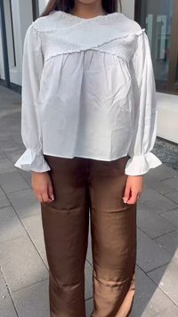 Blusa blanca de manga larga con detalle de smocking texturizado en el escote, combinada con pantalones anchos oscuros de color marrón brillante.
