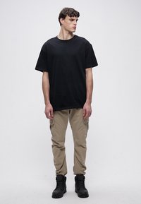 Juodas medvilninis marškinėlis laisvo silueto, derinamas su šviesių chaki spalvos krovinių tipo kelnėmis su šoninėmis kišenėmis ir juodais kariniais batais, suteikiančiais utilitarinį stilių.