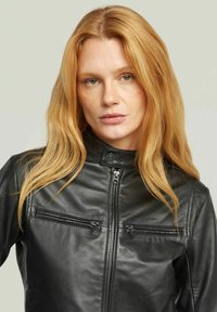 Donna con lunghi capelli rossi che indossa una giacca di pelle nera con tasche con cerniera e un colletto con bottoni a pressione, rivolta in avanti con un'espressione neutra.